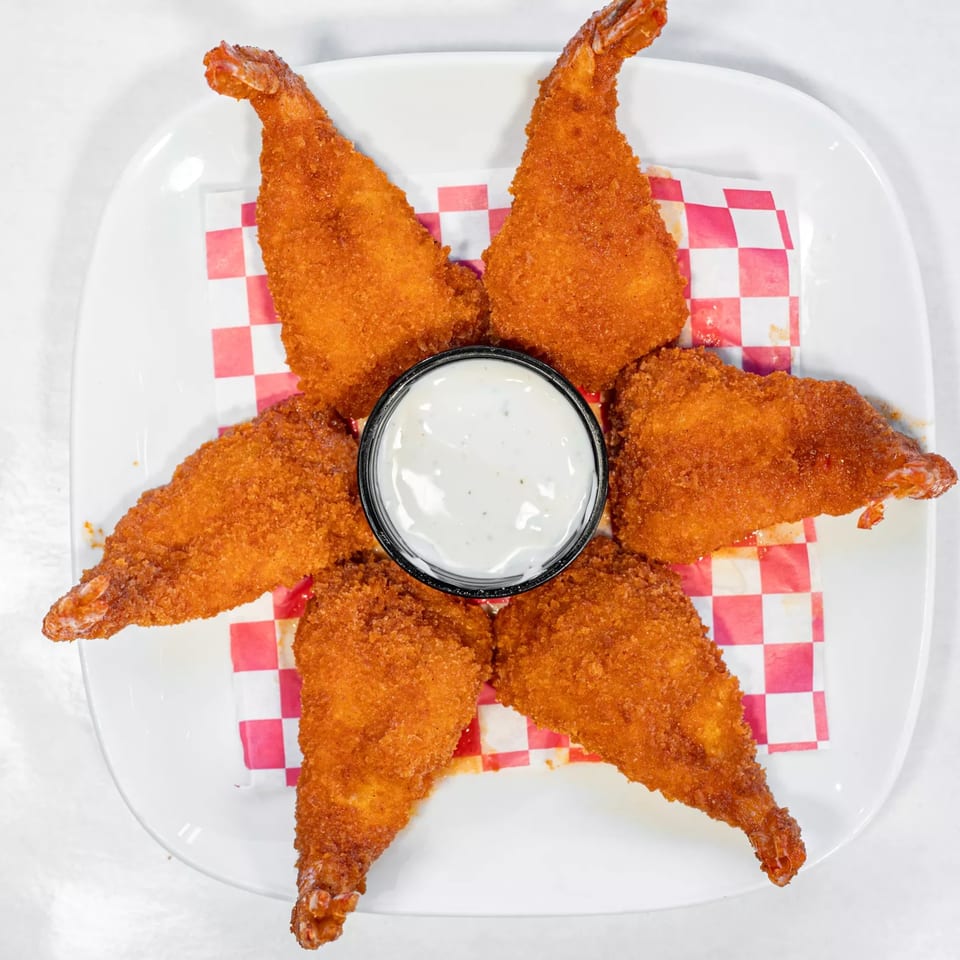 Six crispy fried shrimp arranged around a small cup of white dipping sauce on a white plate.