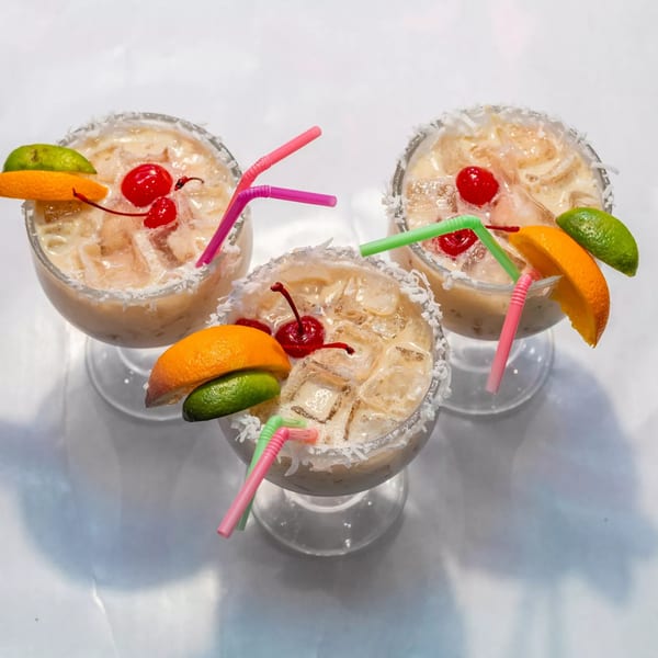 Three creamy cocktails with ice, cherries, orange slices, lime wedges, and colorful straws.
