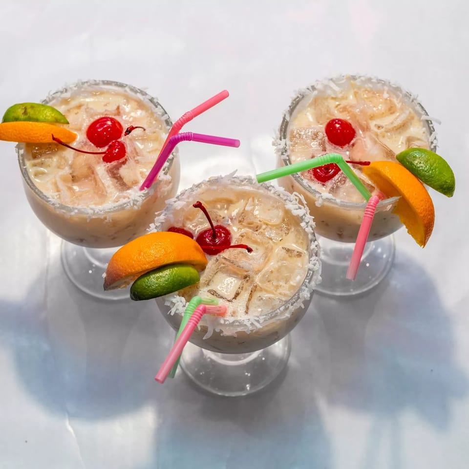 Three creamy cocktails with ice, cherries, orange slices, lime wedges, and colorful straws.