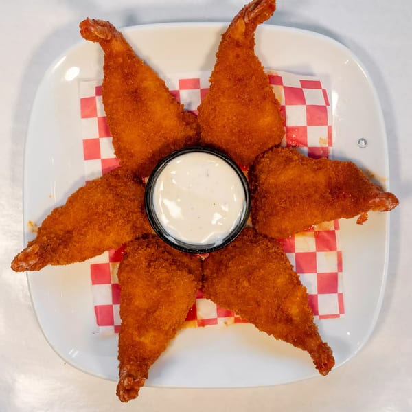 Six crispy fried shrimp arranged around a small cup of white dipping sauce on a checkered paper.