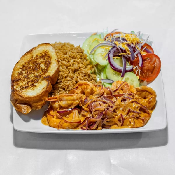 Cajun shrimp with red onions, rice, a side salad with cheese, and toasted bread on a white plate.